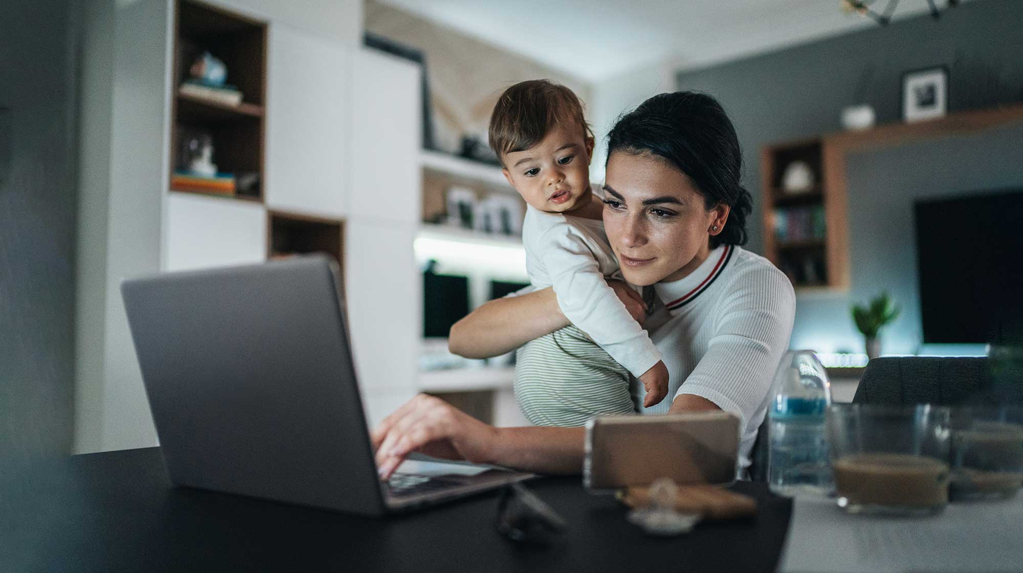 Mother in her home holding a baby while working on laptop